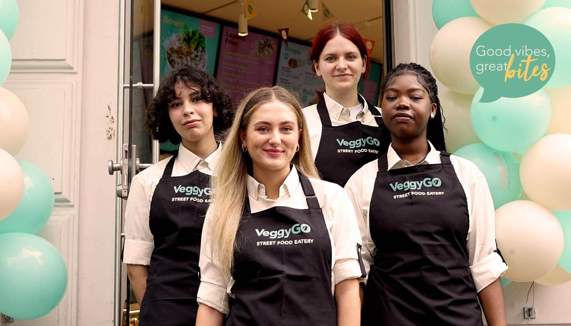 Vier verschiedene Mitarbeiter des Restaurants „Veggygo“, die schwarze Schürzen tragen, lächeln.